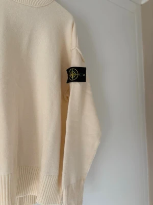 Beige stickad tröja från Stone Island - Snygg beige stickad tröja från Stone Island med klassisk rund halsringning och ribbade muddar. Tröjan har den ikoniska Stone Island-loggan på ärmen och är tillverkad i mjukt material som känns skönt mot huden.