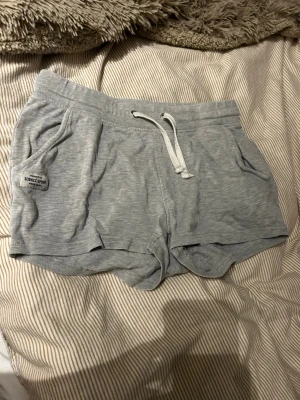 Grå mjuka shorts med snörning - Säljer ett par ljusgrå mjuka shorts från Iconic Spirit. De har snörning i midjan, två fickor fram och en avslappnad passform. Perfekta för chill dagar eller träning. Materialet känns som bomull och de har en enkel, sportig look. De är låg midjade och sitter tajt runt kroppen.
