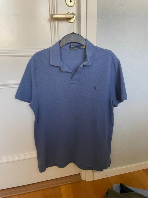 Blå pikétröja från Polo Ralph Lauren - Klassisk blå pikétröja från Polo Ralph Lauren med korta ärmar och broderad logga på bröstet. Tröjan har en mjuk krage och är tillverkad i bomull för en bekväm känsla. Perfekt till jeans eller shorts för en avslappnad stil.