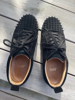 Svarta Spikes Christian Louboutin - Unika svarta sneakers från Christian Louboutin med mattsvarta nitar över tån, mockadetaljer och svarta skosnören. Insidan är i ljust läder och sulan är låg. Perfekta för dig som vill sticka ut med edgy stil.