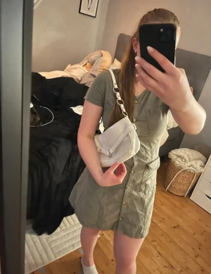 Olivgrön kort klänning med knappar - Säljer en olivgrön kort klänning från H&M med v-ringning och knappar hela vägen framtill. Klänningen har två bröstfickor och två fickor vid höften, samt korta ärmar. Perfekt för dig som gillar en enkel och stilren look. Fin grön klänning med knappar. Kort men inte för kort. Lite för liten för mig därför är det lite mellanrum vid knappar eller så. Men super fin och snygg. Den sitter bra på och har en snygg passform. Perfekt tills sommaren. Den är i storlek 170 men passar dam i S och XS. 