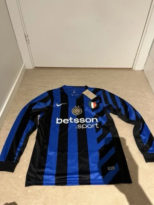 Inter Milan långärmad matchtröja Nike - Snygg långärmad matchtröja från Inter Milan i blått och svart med ränder. Tröjan har klubbmärke, Nike-logga och sponsortryck på bröstet samt italiensk mästerskapsbadge. Tillverkad i ett lätt och ventilerande material, perfekt för fotbollsträning eller match.