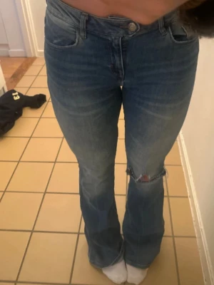 Blå bootcut jeans med slitning - Säljer ett par blå bootcut jeans med slitning vid knät. Jeansen har klassisk femficksmodell, normal midja och är tillverkade i denim. Perfekta för dig som gillar en avslappnad men trendig look.