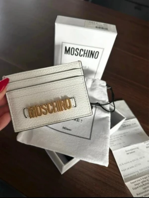 Vit kortfodral från Moschino - Stilrent vitt kortfodral från Moschino i äkta läder med guldfärgad logotyp framtill. Smidig och tunn design med plats för flera kort. Kommer med originalförpackning, dustbag och certifikat. Perfekt accessoar för dig som gillar exklusiva detaljer.