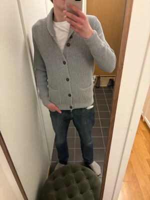 Ljusgrå stickad kofta med sjalkrage - Säljer en ljusgrå stickad kofta med sjalkrage och bruna knappar. Koftan har två framfickor och ribbade muddar vid ärmslut och nederkant. Perfekt för lager på lager och chill stil.