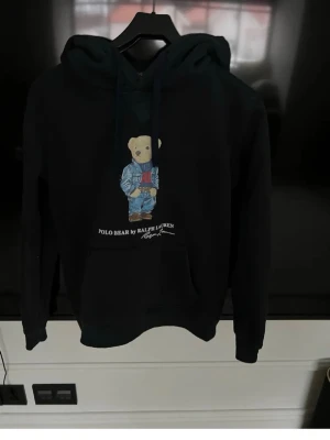 Polo Bear Ralph Lauren - Mörkblå hoodie från Ralph Lauren med Polo Bear-tryck framtill. Klassisk huva. Snygga ikoniska björnen i jeans och RL-tröja. Polo Ralph Lauren