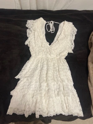 Studentklänning - Supersöt vit kortklänning i spets med volangdetaljer och omlott-v-ringning. Klänningen har knyt i nacken och är ärmlös med romantisk känsla. Perfekt för sommarens alla tillfällen och ger en fräsch och feminin look.