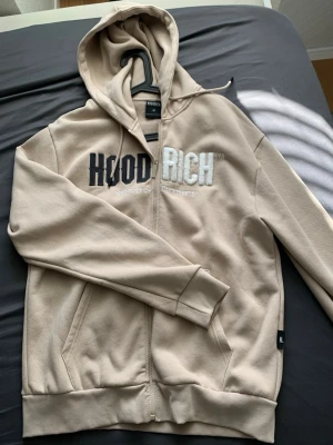 Beige zip hoodie från Hoodrich - Snygg beige hoodie med dragkedja från Hoodrich. Stor broderad logga i svart och vitt på bröstet. Utrustad med huva och dragsko samt två fickor framtill. Tillverkad i mjukt bomullsmaterial för skön känsla. Perfekt för en avslappnad streetwear-look.