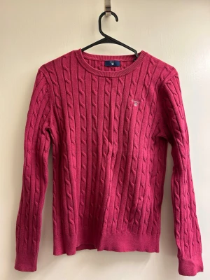 Rosa kabelstickad tröja från Gant - Kabelstickad tröja i en snygg rosa färg från Gant. Tröjan har rund halsringning, långa ärmar och ribbade muddar. Liten broderad Gant-logga på bröstet. Perfekt för lager på lager och en klassisk look.