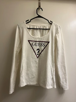 Vit långärmad topp från Guess - Snygg vit långärmad topp från Guess med det klassiska triangellogotypen och texten 'GUESS?' framtill. Tillverkad i mjuk bomull med rund halsringning. Perfekt för en clean och stilren look.