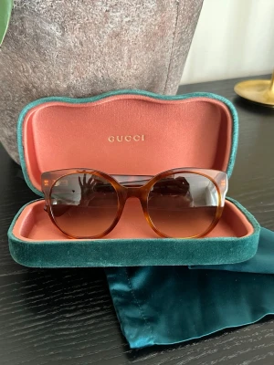 Bruna solglasögon från Gucci - Snygga solglasögon från Gucci med brunsköldpaddsmönstrad båge och klassisk rundad form. Sidorna pryds av den ikoniska guldiga Gucci-loggan. Kommer med ett lyxigt fodral i grön sammet och rosa insida.     Modell: GG0355S Färgkod: 012 Storlek: 54-22 Skalmängd: 140   Välomhändertagna. Finns små repor på högra glaset. 