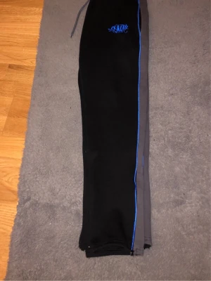 Svarta mjukisbyxor med blå detaljer - Säljer ett par svarta mjukisbyxor från Stay med blå piping längs sidorna och broderad logga på höften. Byxorna har resår i midjan med dragsko och dragkedjor vid bensluten för extra stil. Perfekta för chill eller streetwear. Xs men passar s. Byte funkar också 