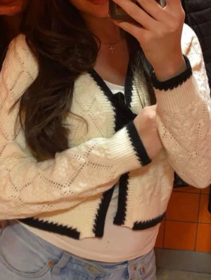 Vit kofta med svarta detaljer - Snygg vit kofta med svarta kanter och diskret mönsterstickning. Kortare modell med långa ärmar och ribbade muddar. Perfekt att slänga över en topp för en trendig look.