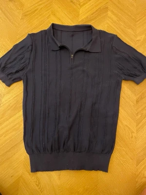 Mörkblå stickad pikétröja - Snygg mörkblå stickad pikétröja med kort ärm och dragkedja vid halsen. Tröjan har diskreta vertikala ränder i strukturen och ribbad mudd nedtill. Perfekt för en stilren och avslappnad look.