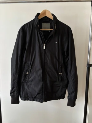 Svart vindjacka från J.Lindeberg - Stilren svart vindjacka från J.Lindeberg med hög krage, dragkedja framtill och två fickor med knappstängning. Jackan har diskret logga på bröstet och ribbade muddar vid ärmslut och nederkant. Tillverkad i lätt och tåligt material.