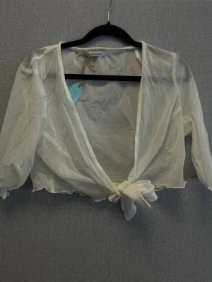 Vit transparent knytblus från Goldmann - Snygg vit transparent blus från Goldmann med knyt framtill och långa ärmar. Blusen har en djup v-ringning och är gjord i ett tunt, lätt material som ger en luftig känsla. Perfekt för lager på lager och trendiga outfits.