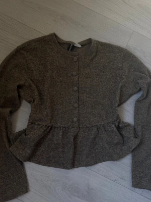 Brun topp från H&M - Mysig brun topp kofta från H&M med peplumdetalj i midjan och rund halsringning. Koftan har långa ärmar och knappar framtill i samma färg som plagget. Perfekt för lager på lager och enkel att styla till olika outfits. Känns som storleken xs/s men den är i medium.