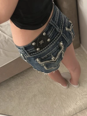 Mörkblå jeansshorts från Miss Me - Säljer ett par mörkblå jeansshorts från Miss Me med dekorativa sömmar, slitna kanter och snygga detaljer med nitar och broderi på bakfickorna. Klassisk femficksmodell och låg midja. Perfekta för sommaren! 36cm rakt över midjan❤️