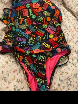Färgglad Nike baddräkt  - Säljer en supercool baddräkt från Nike med färgglatt grafiskt tryck och text som 'Just Do It' och smileys. Svart bas med neonfärger och rosa insida. Baddräkten har tunna axelband som korsas i ryggen och är perfekt för dig som vill sticka ut på stranden eller i poolen.