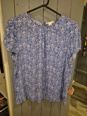 Blå blommig blus med kort ärm - Blå blus med vitt blommönster och rund halsringning. Blusen har korta ärmar och knappdetalj i nacken. Tillverkad i ett lätt och luftigt tyg som ger en avslappnad känsla. Perfekt för dig som gillar mönstrade plagg och vill ha något bekvämt och snyggt. Aldrig använd.