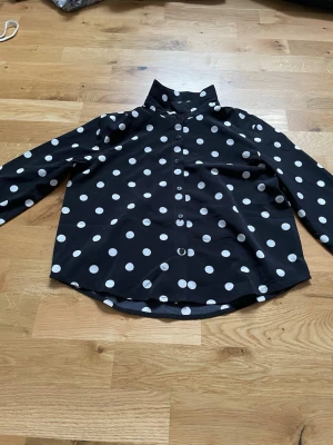 Svart prickig skjorta från KappAhl - Snygg svart skjorta från KappAhl med stora vita prickar och klassisk krage. Skjortan har långa ärmar och knäppning framtill. Perfekt för dig som vill sticka ut med ett coolt mönster och en trendig look.