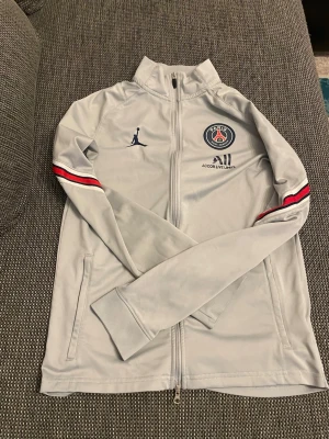 PSG x Jordan ljusgrå träningsjacka - Ljusgrå träningsjacka från PSG och Jordan med hel dragkedja, ståkrage och fickor. Jackan har röda och svarta ränder på ärmarna samt PSG-logga och Jumpman-logga på bröstet. Tillverkad i ett mjukt, stretchigt material som passar perfekt till träning.