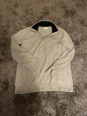 Ljusgrå halvzip tröja från Oscar Jacobson - Snygg ljusgrå tröja från Oscar Jacobson med halvzip och hög krage. Tröjan har ribbade muddar vid ärmslut och nederkant samt svart insida på kragen. Perfekt för lager på lager och enkel att matcha.