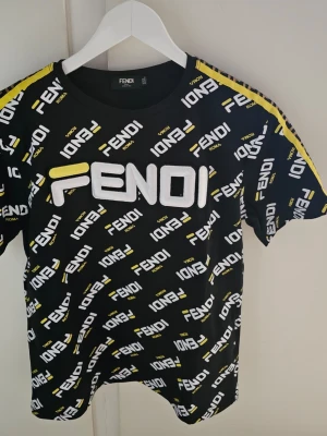 Svart Fendi t-shirt med logotypmönster - Snygg svart t-shirt från Fendi med stort broderat FENDI-tryck på bröstet och all-over logotypmönster i vitt och gult. T-shirten har gula och vita detaljer längs axlarna och är kortärmad. Perfekt för dig som vill sticka ut med en exklusiv streetstyle-look.