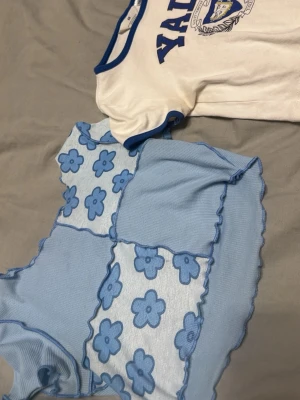 Vit Yale t-shirt och blå blommig topp st. 146 - Säljer en vit t-shirt från H&M med blå Yale-tryck och blå kant vid halsen. Medföljer gör en ljusblå mesh-topp med blommigt mönster och dekorativa overlock-sömmar i blått. Perfekta för en trendig och avslappnad stil. Storlek 146