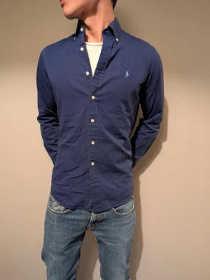 Marinblå skjorta från Ralph Lauren - Klassisk marinblå skjorta från Ralph Lauren med broderad logga på bröstet. Skjortan har knappar hela vägen, button-down krage och långa ärmar. Tillverkad i mjuk bomull som känns skön mot huden. Perfekt för en clean och stilren look.