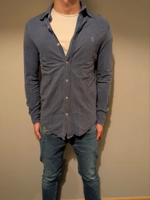 Blå skjorta från Ralph Lauren - Säljer en klassisk blå skjorta från Ralph Lauren med lång ärm och knappar framtill. Skjortan har den ikoniska broderade loggan på bröstet och är tillverkad i mjuk bomull. Perfekt till jeans eller chinos för en clean look.