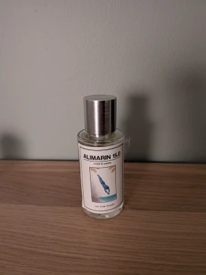 Alimarin 15.0 parfymflaska - En stilren parfymflaska från Alimarin med namnet 15.0. Flaskan är genomskinlig med en metallfärgad kork och har en vit etikett med blått motiv. Perfekt för dig som gillar minimalistisk design och fräscha dofter. Helt ny, endast några få testsprays använda☺️ Inspirerad av Lv Imagination😁