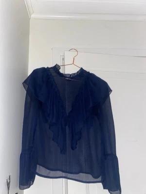 Mörkblå volangblus med transparent tyg - Snygg mörkblå blus i transparent material med volangdetaljer framtill och vid kragen. Blusen har lång ärm och en lätt rynkad halsringning som ger en romantisk känsla. Perfekt att styla med jeans eller kjol för en trendig look, köpt från ett UF företag så vet inte vart den kommer från ursprungligen, använd en gång!