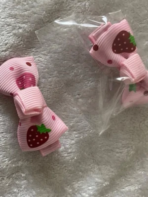 Rosa hårspännen med jordgubbsmotiv - Säljer två söta hårspännen i rosa tyg med jordgubbsmotiv och små prickar. Spännena har en klassisk rosettform och fästs enkelt med metallclips på baksidan. Perfekta för att piffa upp frisyren med en gullig detalj. 25kr/par 