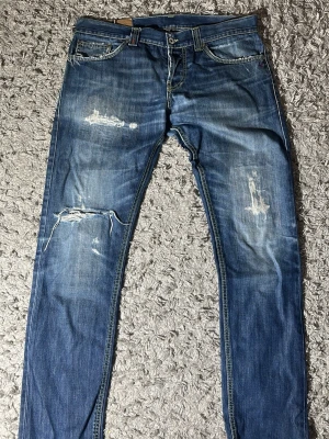 Blå dondup jeans med schyssta slitningar skinny fit - Säljer ett par blå jeans med slitningar och raka ben. Jeansen har en klassisk femficksmodell och tydliga slitdetaljer på lår och knän för en avslappnad look. Perfekta för dig som gillar en trendig och casual stil.