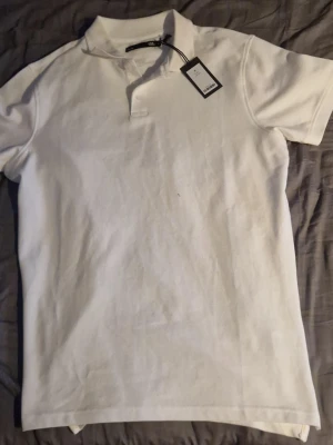 Vit pikétröja från Lager 157 - Enkel och stilren vit pikétröja från Lager 157 med klassisk krage och korta ärmar. Tillverkad i mjuk bomull och har knappar vid halsen för en clean look. Perfekt till jeans eller shorts för en avslappnad stil.