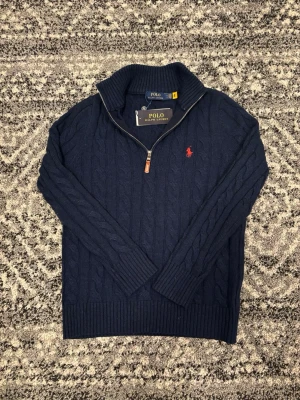 Mörkblå kabelstickad tröja Polo Ralph Lauren - Mörkblå kabelstickad tröja från Polo Ralph Lauren med halv dragkedja. Tröjan har det klassiska röda Polo-logot på bröstet. Tröjan är i storleken S. Hör av er vid eventuella frågor eller funderar!!