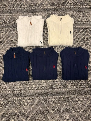 Stickade tröjor från Ralph Lauren - Fem stickade tröjor från Ralph Lauren med kabelstickat mönster och halv dragkedja. Två i vitt och tre i marinblått. Alla med klassiska röda logga. Tröjorna är från storlek S till M. Hör av er vid eventuella frågor eller funderingar!!