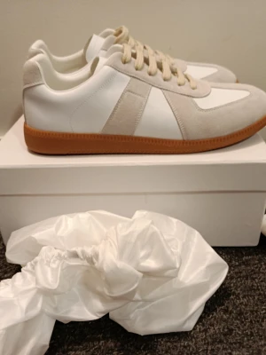 Maison Margiela vita sneakers - Stilrena vita sneakers från Maison Margiela med beige mockadetaljer och klassisk gummisula i brunt. Skorna har en retroinspirerad design med vita skosnören och diskreta sömmar. Perfekta för dig som gillar minimalistisk och trendig stil.