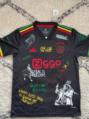 Ajax x Bob Marley limited edition tröja - Unik svart Ajax x Bob Marley limited edition tröja från Adidas med färgglada detaljer i rött, gult och grönt på axlarna. Tryck med Ziggo-logga, Bob Marley-motiv och inspirerande citat. Perfekt för dig som älskar fotboll och reggae.