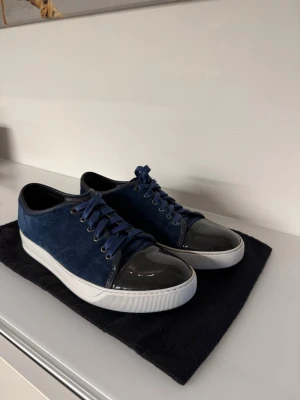 Lanvin - Snygga sneakers i mörkblå mocka med matchande blå skosnören och vit sula. Tåpartiet har en unik svart lackdetalj som ger extra edge. Perfekta för dig som vill ha en stilren men ändå cool look. Passar till jeans eller chinos för en avslappnad vibe. Storlek UK 7. Vilket ska motsvara 41 i vanlig storlek men passar mig perfekt som brukar ha storlek 42-43. Hör gärna av er vid frågor:) 