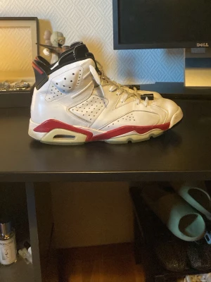 Air Jordan 6 2010 Retro Vit/Röd - Säljer ett par Air Jordan 6 från 2010 Retro sneakers i vitt läder med röda och svarta detaljer. Skorna har perforerade paneler, synlig Air-sula och klassisk hög siluett. Perfekt för dig som gillar ikoniska basketskor och streetwear.
