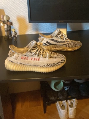 Yeezy Boost 350 Zebra sneakers - Säljer ett par Adidas Yeezy Boost 350 V2 'Zebra' sneakers med svartvitt zebramönster, vit sula och röda 'SPLY-350' detaljer på sidan. Ovandelen är i stickad mesh och skorna har klassisk låg siluett med snörning. Perfekta för dig som gillar streetwear och unik stil. Defekt synd på bild. Storlek 43 1/3