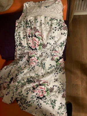 Vit blommig klänning med spetsdetalj - Vit ärmlös klänning med blommigt mönster i rosa, grönt och svart. Överdelen har en snygg spetsdetalj som ger en romantisk touch. Kjolen är lätt utsvängd och klänningen är perfekt för sommardagar. Tillverkad i ett mjukt bomullsmaterial.