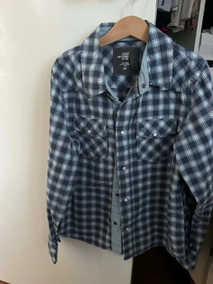 Blårutig skjorta från H&M - Snygg blårutig skjorta från H&M med klassisk krage och två bröstfickor med knappar. Skjortan har långa ärmar och stängs med knappar framtill. Perfekt för en avslappnad och trendig look.