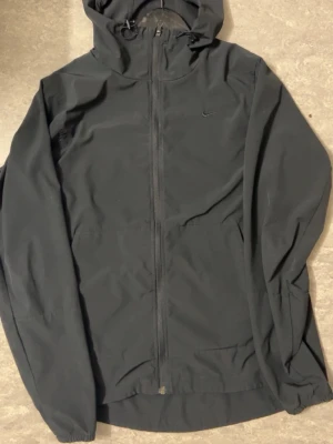 Svart vindjacka från Nike med huva - Snygg svart vindjacka från Nike med hel dragkedja och huva. Jackan har minimalistisk design med diskret Nike-logga på bröstet och elastiska muddar vid ärmsluten. Tillverkad i lätt och tåligt material, perfekt för blåsiga dagar.