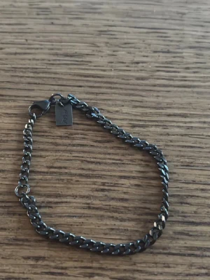 Svart kedjearmband i metall - Snyggt svart kedjearmband i metall med en liten rektangulär bricka nära låset. Armbandet har en klassisk karbinhake och stilren design som passar till många olika looks. Perfekt för dig som gillar minimalistiska accessoarer.