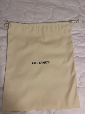 Beige dustbag från Axel Arigato - Säljer en beige dustbag från Axel Arigato med dragsko upptill. Perfekt för att förvara skor eller accessoarer. Dustbagen är tillverkad i ett mjukt tygmaterial och har enkel, stilren design med logga tryckt i svart.