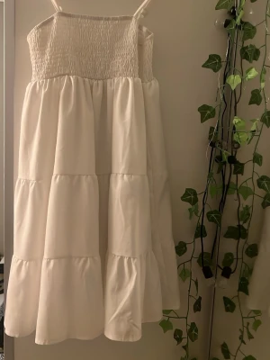 Vit sommarklänning med volanger - Luftig vit klänning med smock upptill och tunna axelband. Kjolen har flera volanglager som ger en romantisk känsla. Perfekt för varma dagar och enkel att styla med accessoarer.