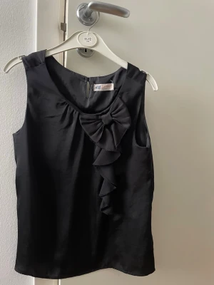 Svart blus från H&M - Snygg svart blus från H&M i ett glansigt material med volangdetalj framtill. Modellen är ärmlös och har en rund halsringning. Perfekt att styla med jeans eller kjol för en trendig look. Väldigt elegant, ren och fin. 